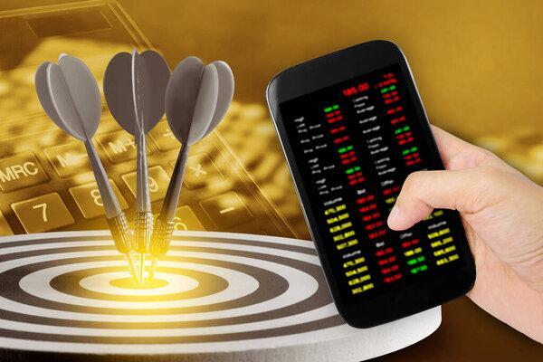 Hand holdind smartphone and stock charts screen with  Circular target marked  numbers and dart with credit card and caculater background. An idea of business targets, audience, market, group, analysis, risk, rate, area, value .